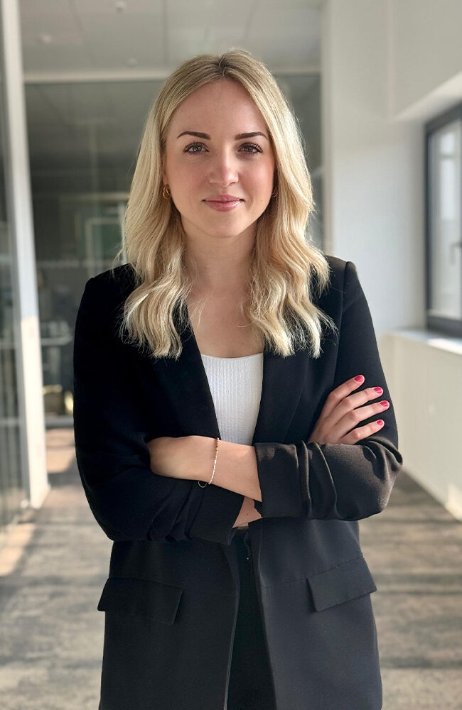 Karriere 5 Anne - Junior HR Manager | Eine Frau mit langen blonden Haaren steht in einem modernen Büro. Sie trägt ein schwarzes Oberteil und hat die Arme verschränkt.